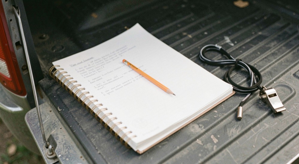 A spiral notebook open on the tailgate of a truck, a pencil lying across the page and a whistle lanyard coiled beside it in flat light