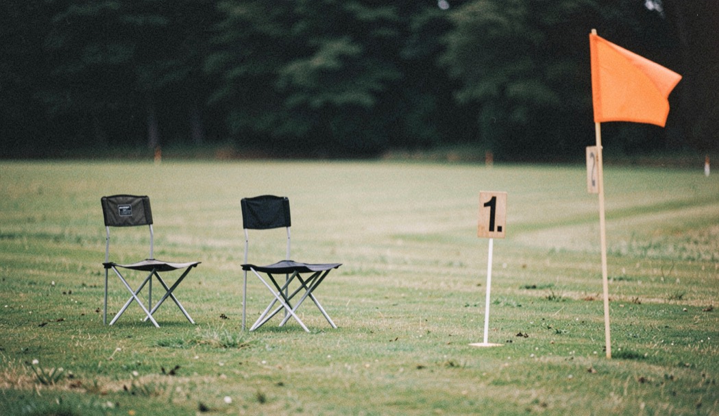 Two folding chairs and a numbered stake at the edge of a mown field, an orange marker flag bent by the wind and a dark treeline behind